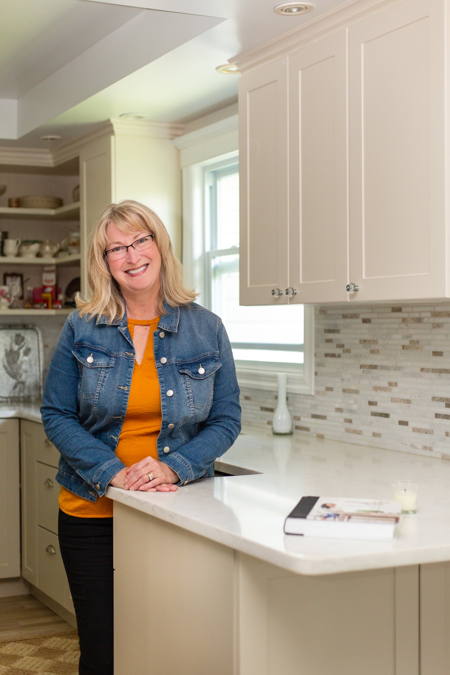 Woman standing in a modern kitchen with white cabinets and a window in the background.